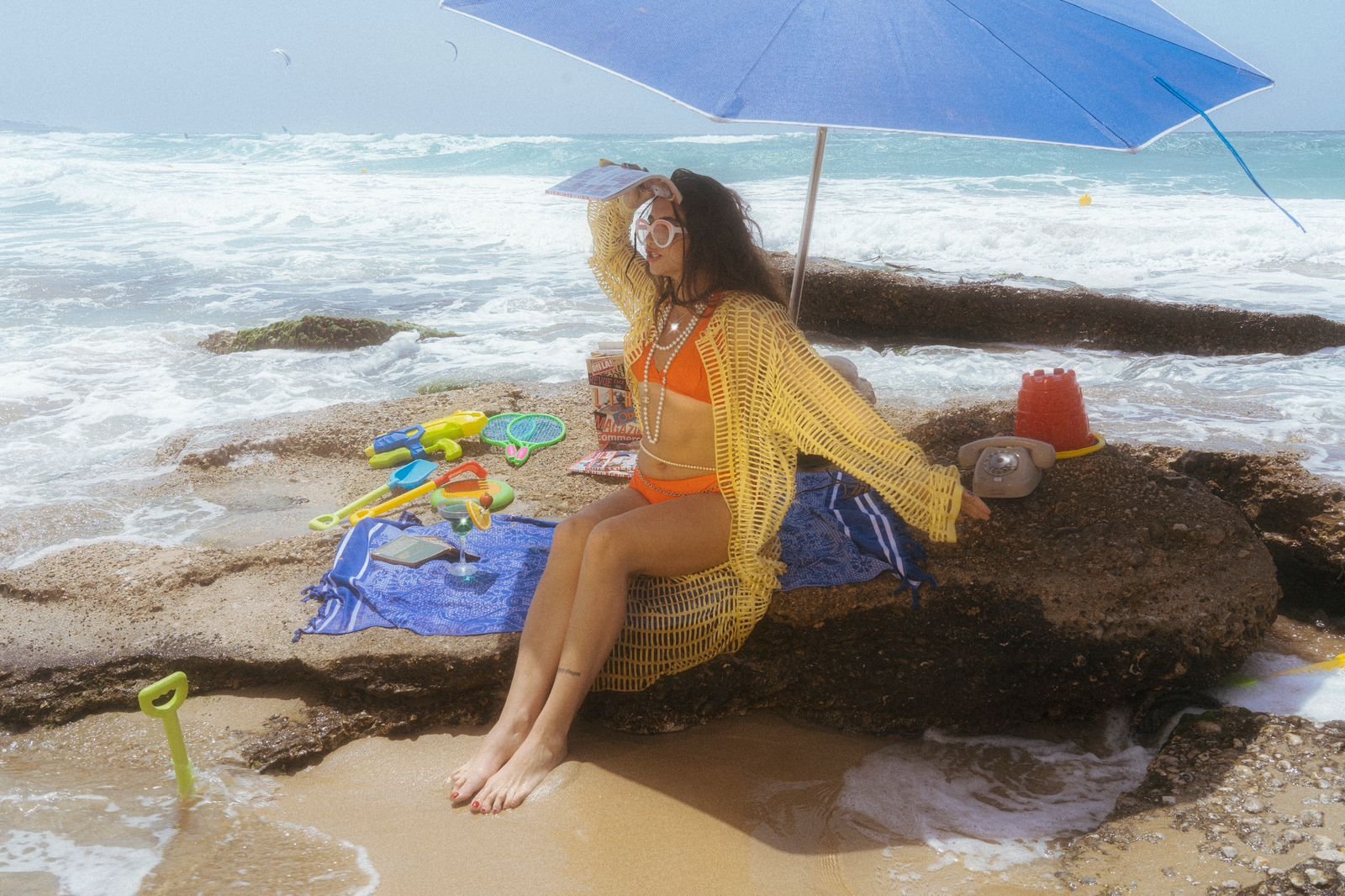 A woman in an orange swimsuit and yellow crochet cover-up sitting on rocks under a blue parasol