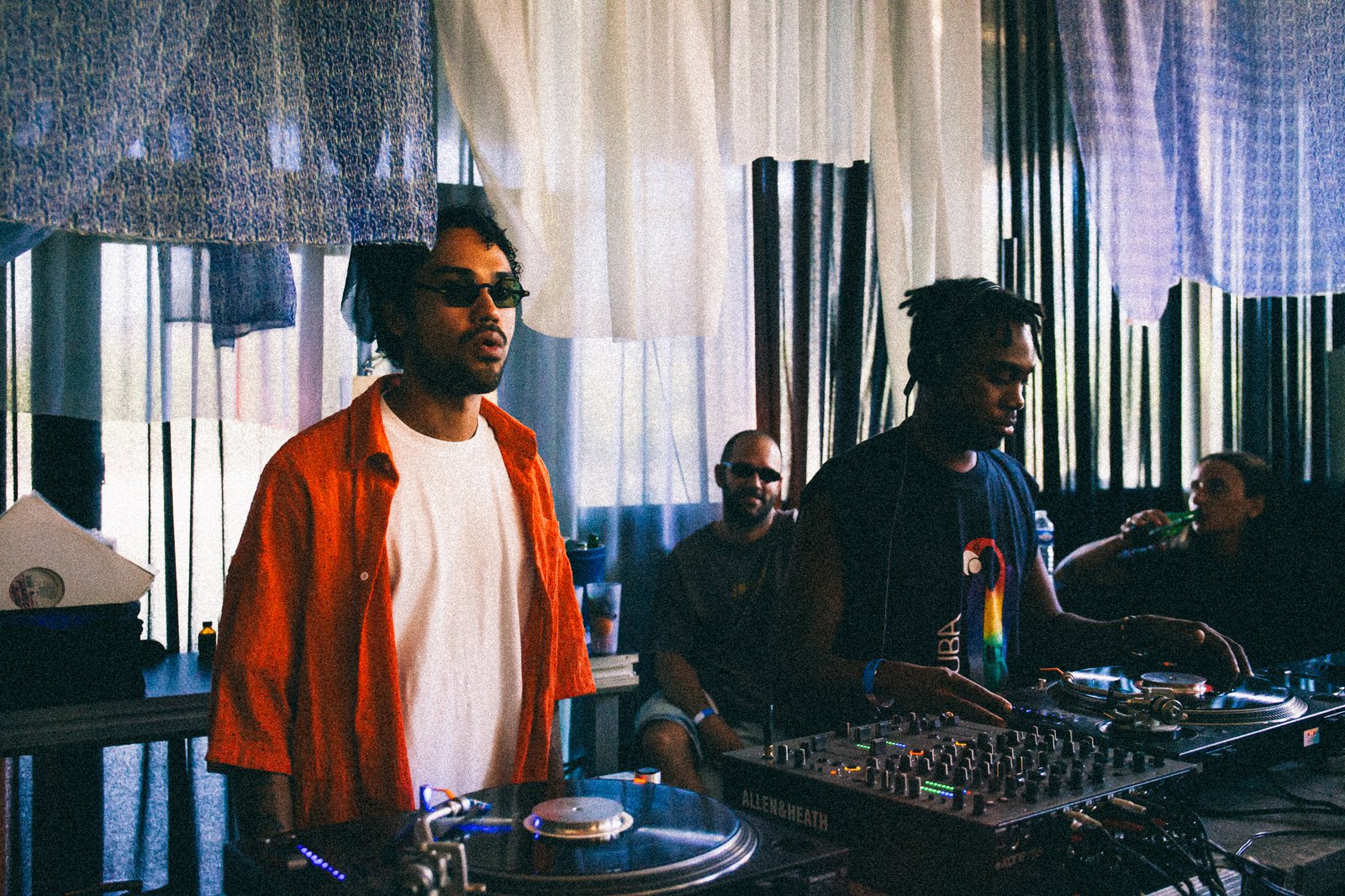 A DJ in an orange shirt at the decks in front of blue and white patterned drapes