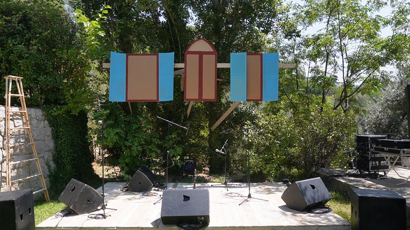 The set by day: painted shutters and an arched frame mounted on a timber beam between the trees, speakers and a ladder on the stage