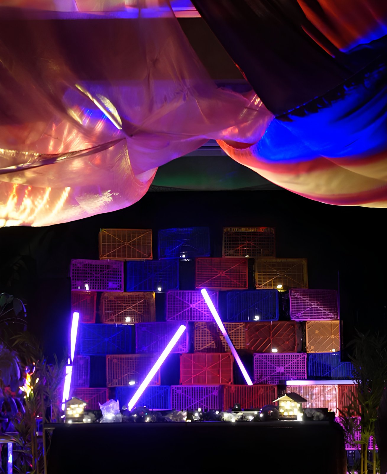 The stage: a wall of stacked wooden crates lit in pink and blue with neon tubes, under a draped fabric canopy