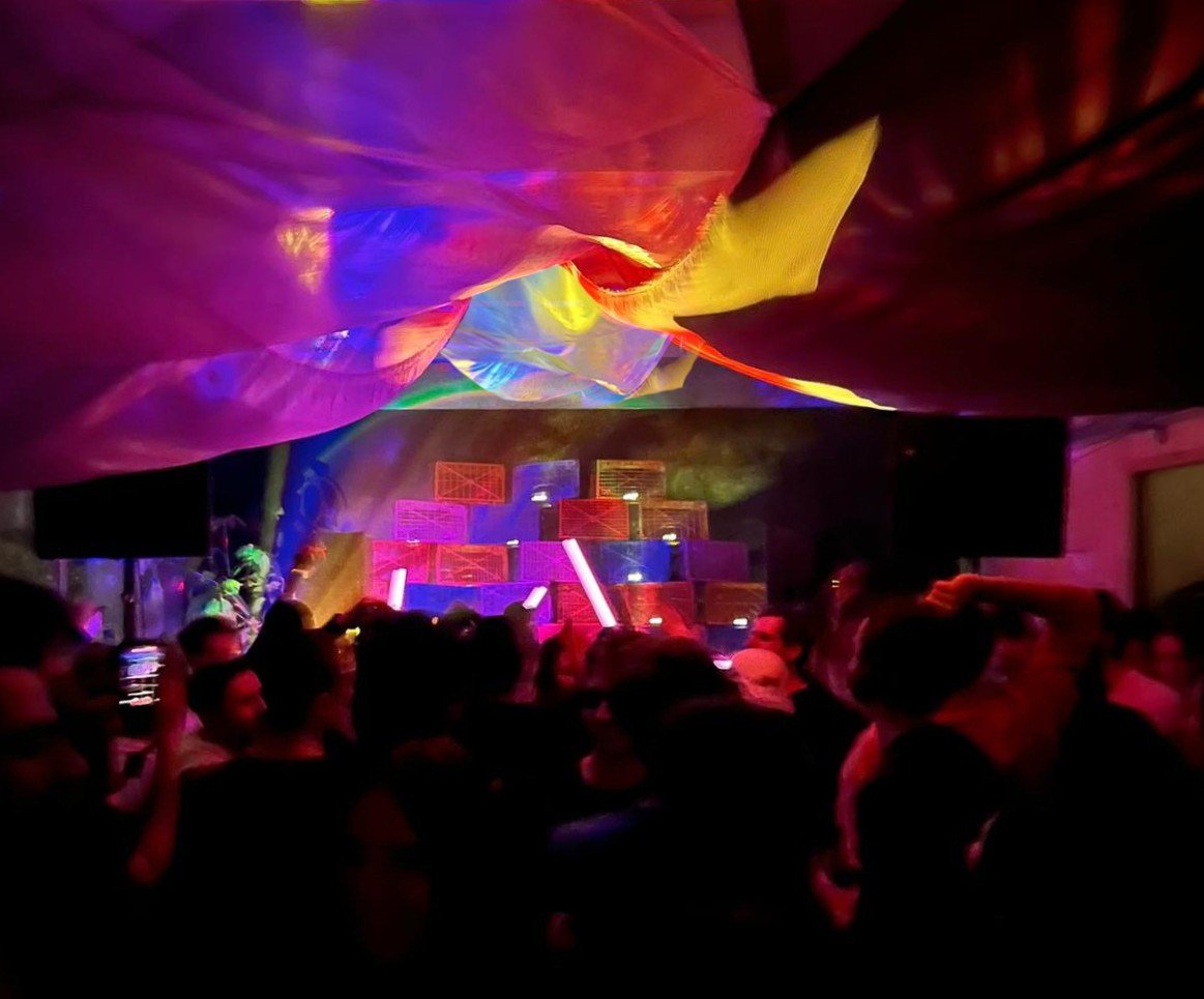 The crowd under the coloured fabric canopy, the crate wall and neon glowing behind
