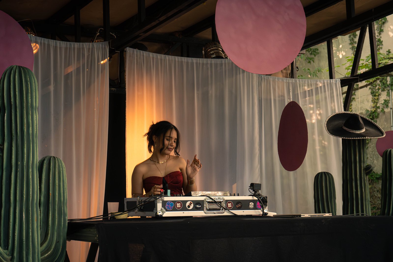 A DJ behind the decks under a wooden pergola, white drapes, plum-coloured discs and tall cacti around her