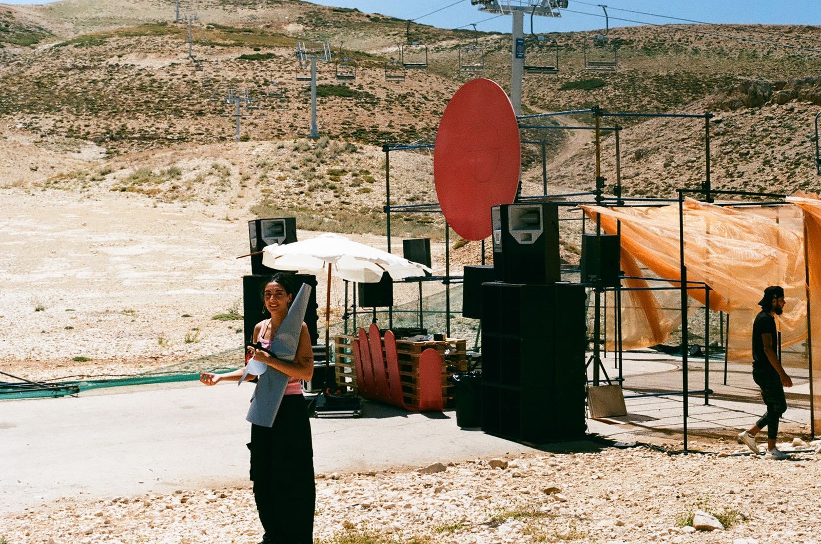 The Microfest stage being built on a bare mountainside: scaffold frames, speaker stacks, a large red disc and a parasol, with a ski lift behind