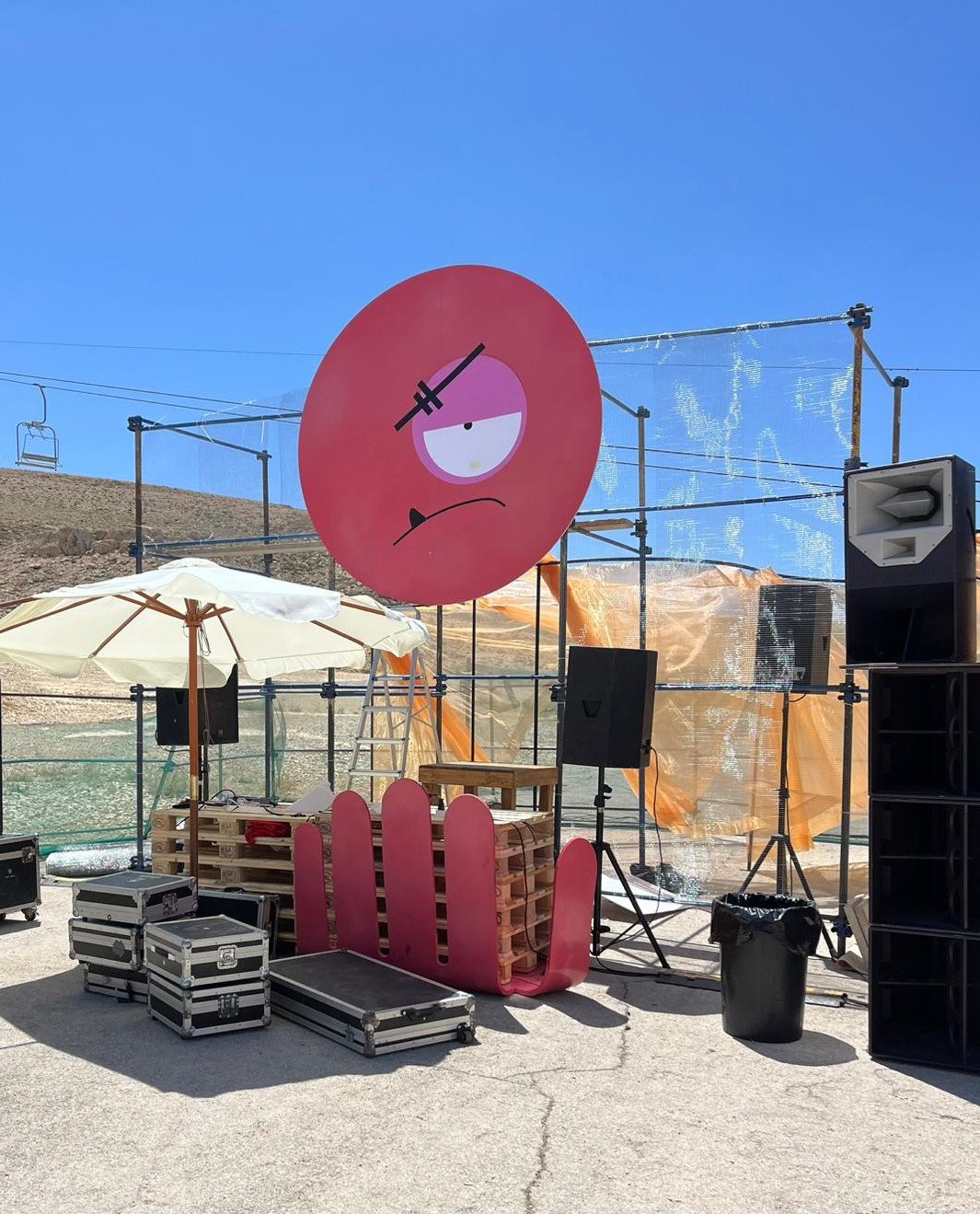 The red disc with a painted face above the stage scaffold, flight cases and a pink hand-shaped cut-out in the foreground