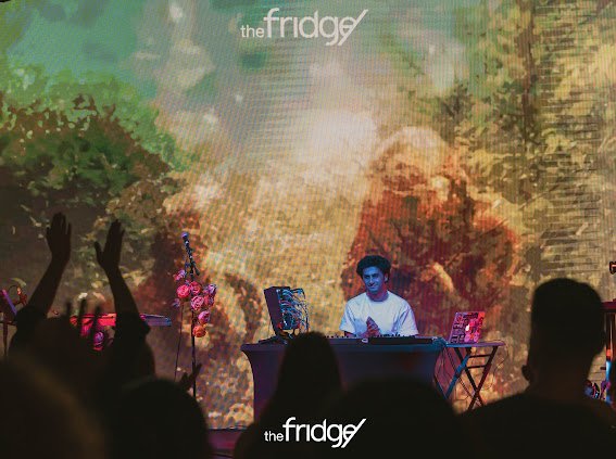 Serge Volant at his desk on stage under The Fridge logo, a warm floral projection filling the wall behind him and the crowd in silhouette